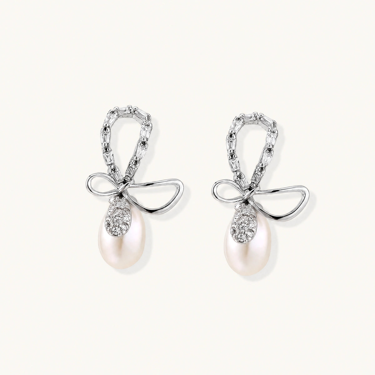 Silver bow-shaped stud earrings with baguette crystals and dangling white freshwater pearls on cream background